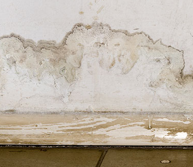 The image shows a damaged interior wall with significant water damage, peeling paint, and discoloration along the baseboard area, indicating moisture infiltration or flooding issues.